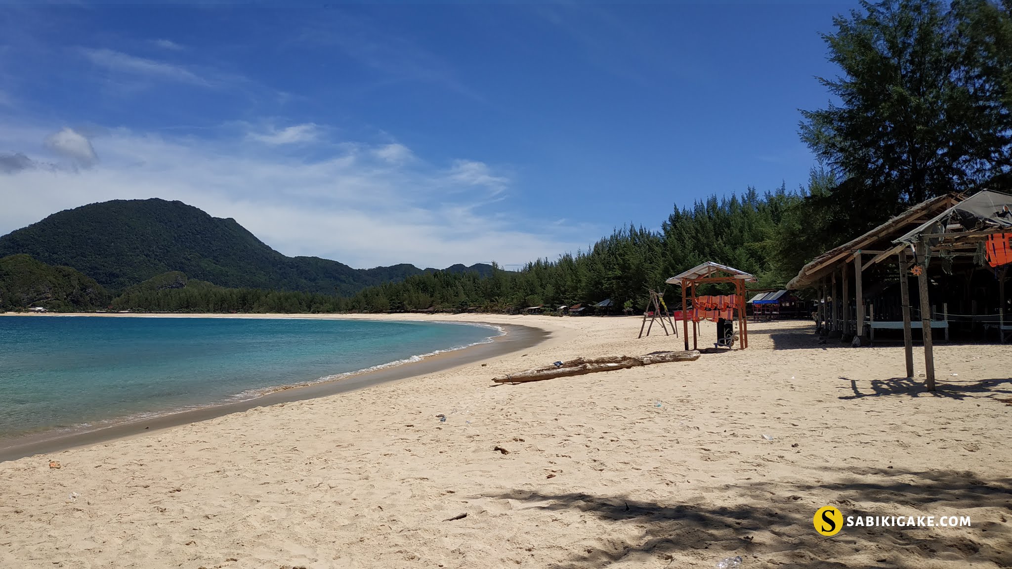 Pemandangan Pantai Lampuuk di Aceh Besar dengan pasir putih dan air laut jernih, dikelilingi pepohonan dan bukit di latar belakang.